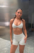 A woman modeling a light gray two-piece athletic set consisting of a halter-style sports bra and matching high-waisted shorts, standing in front of a metallic wall.